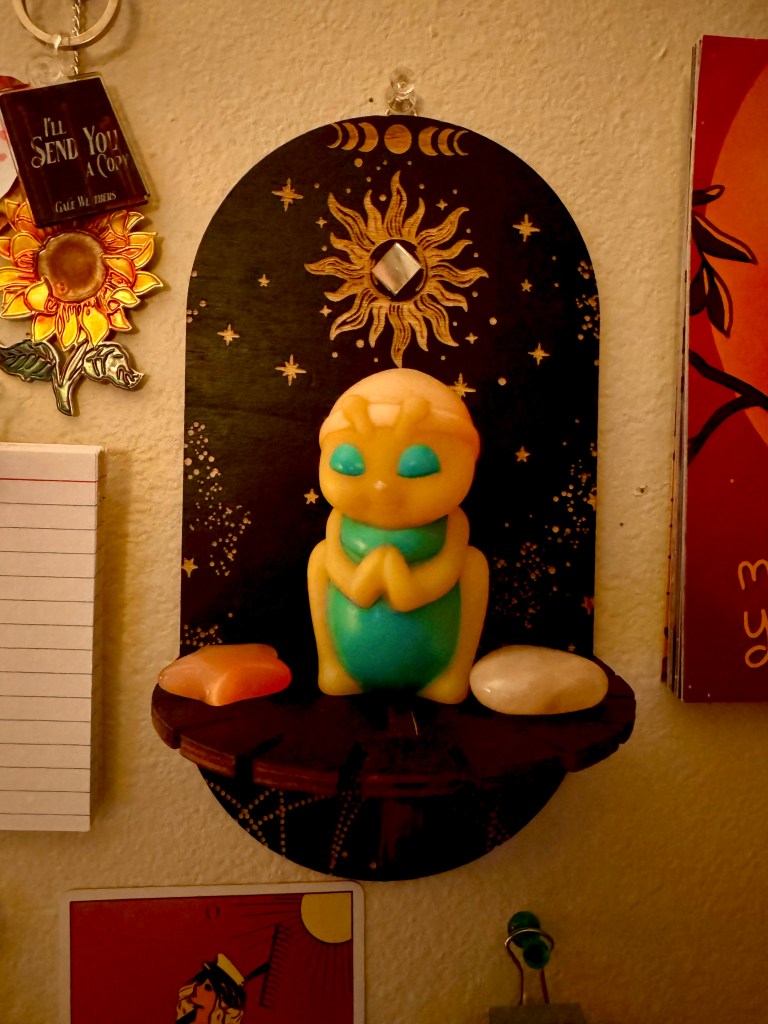 A small hanging shelf, black with gold accents like the moon cycle and a sun and stars, with a small square of mirror glued to the inside of the sun. On the shelf are two rose quartz stones with a praying Glo-Worm toy between them.
